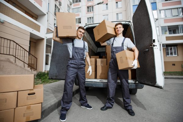 Two young handsome smiling workers wearing uniforms are standing in front of the van full of boxes holding boxes in their hands. The block of flats is in the background. House move, mover service.